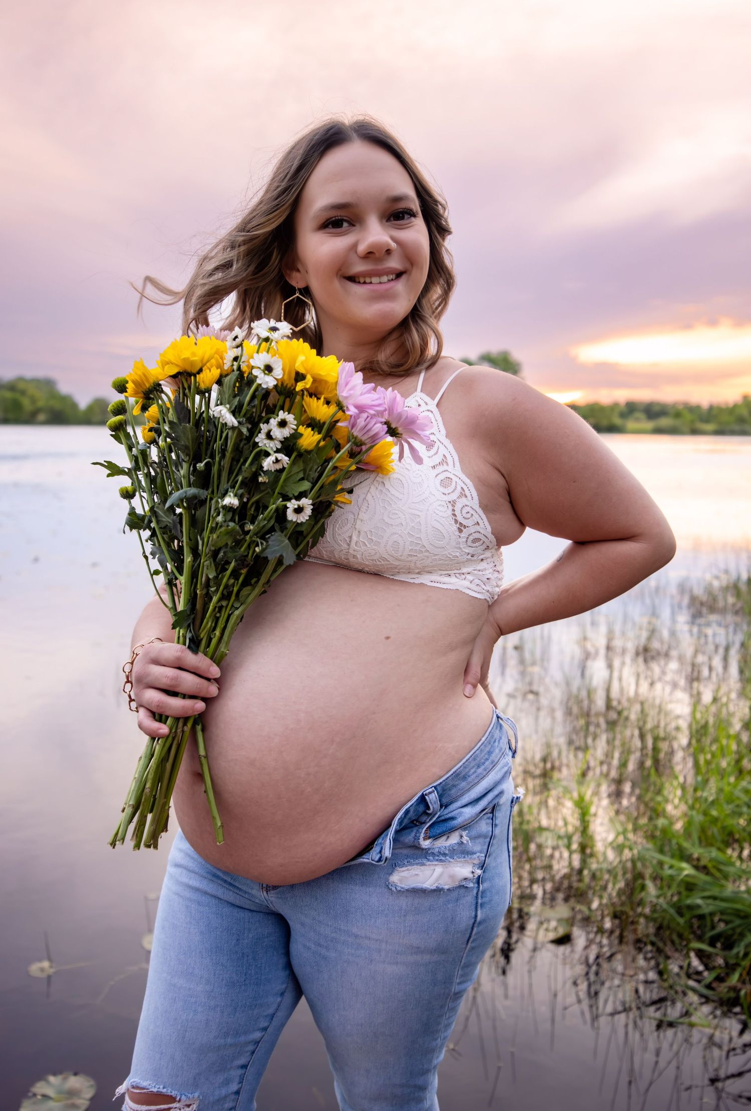 Maternity photo with wildflower bouquet by lake during sunset.
