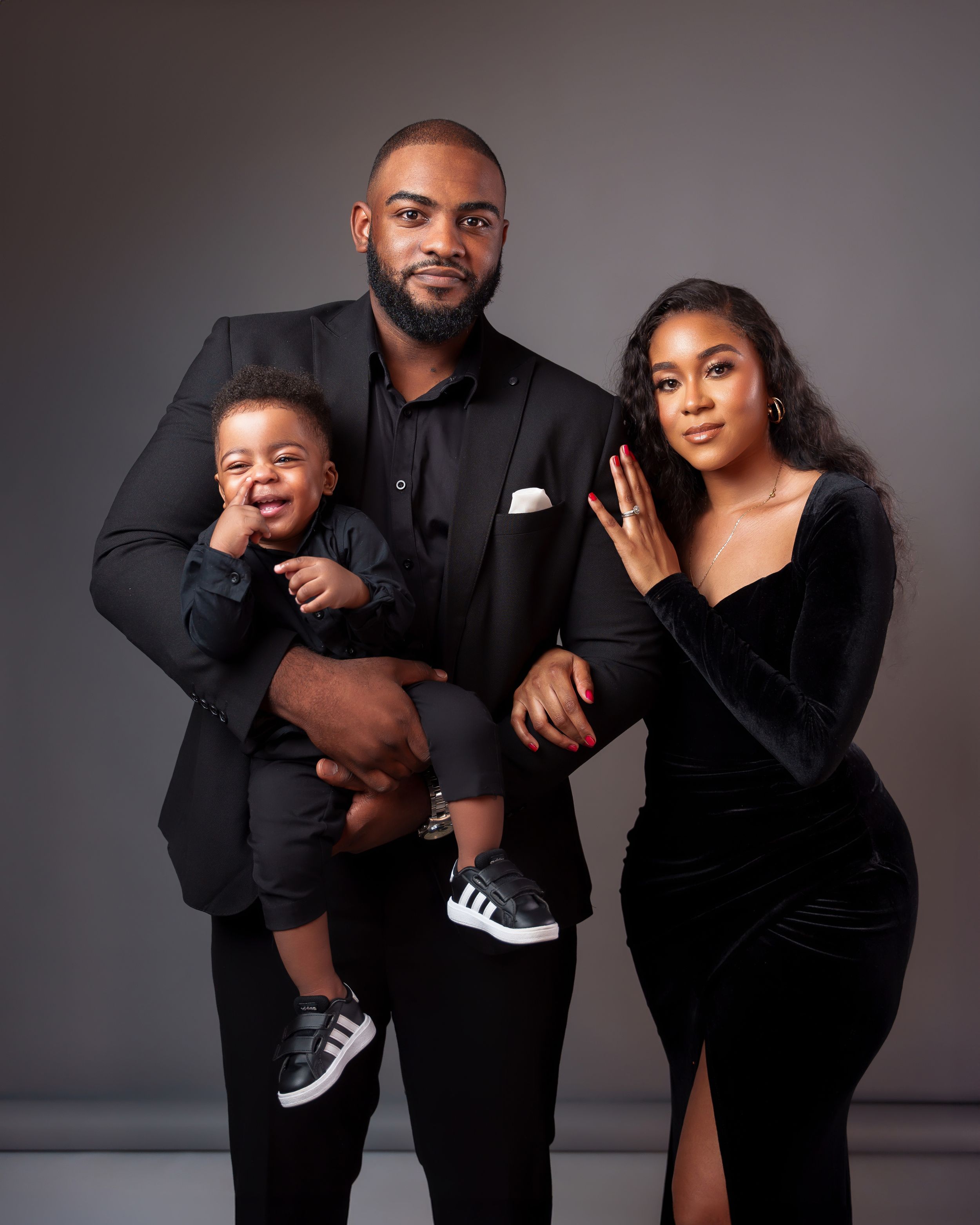 Stylish family portrait with couple in black formal wear and toddler wearing dress shoes captured against dark background.