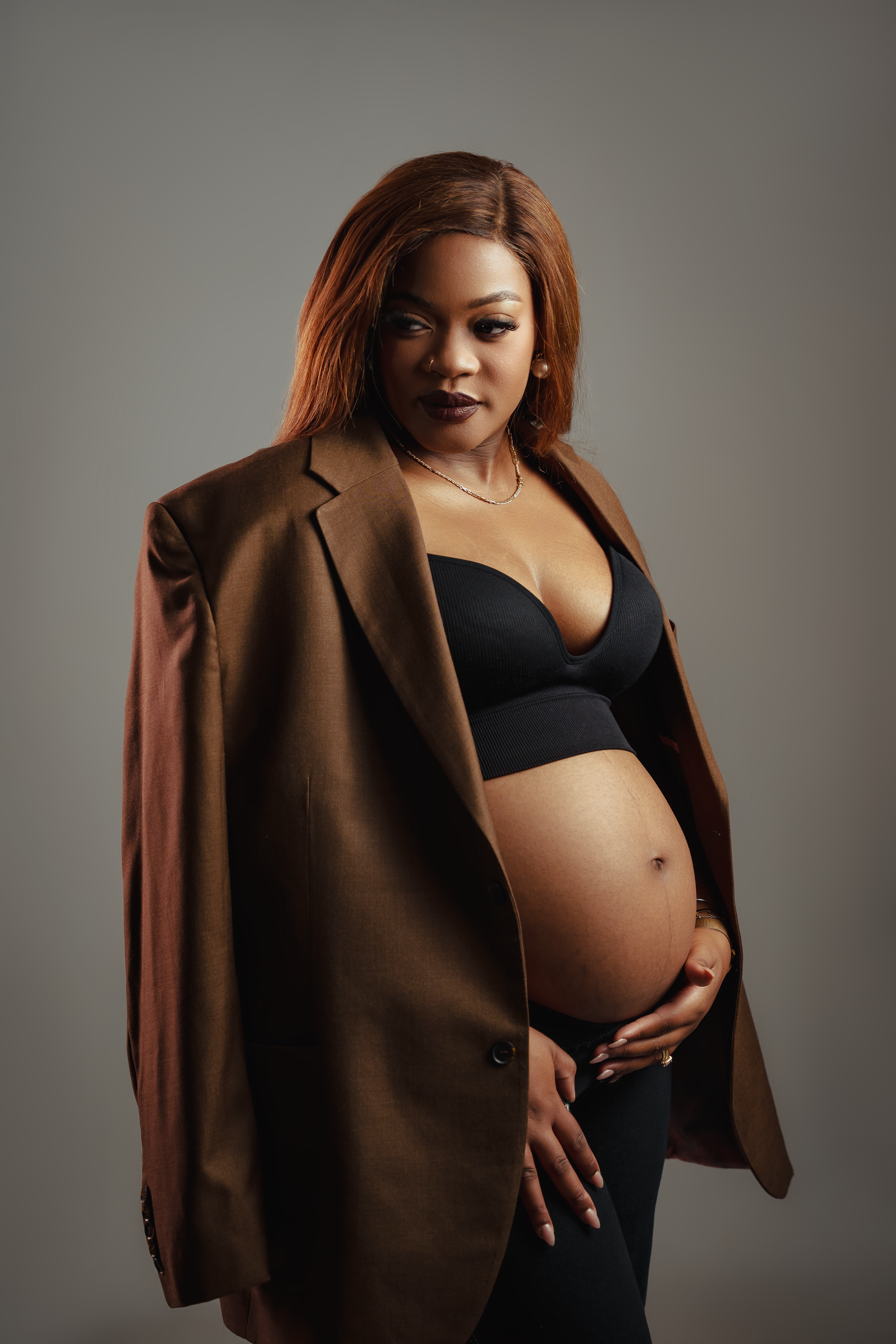 Maternity portrait with brown coat and black outfit against neutral backdrop.