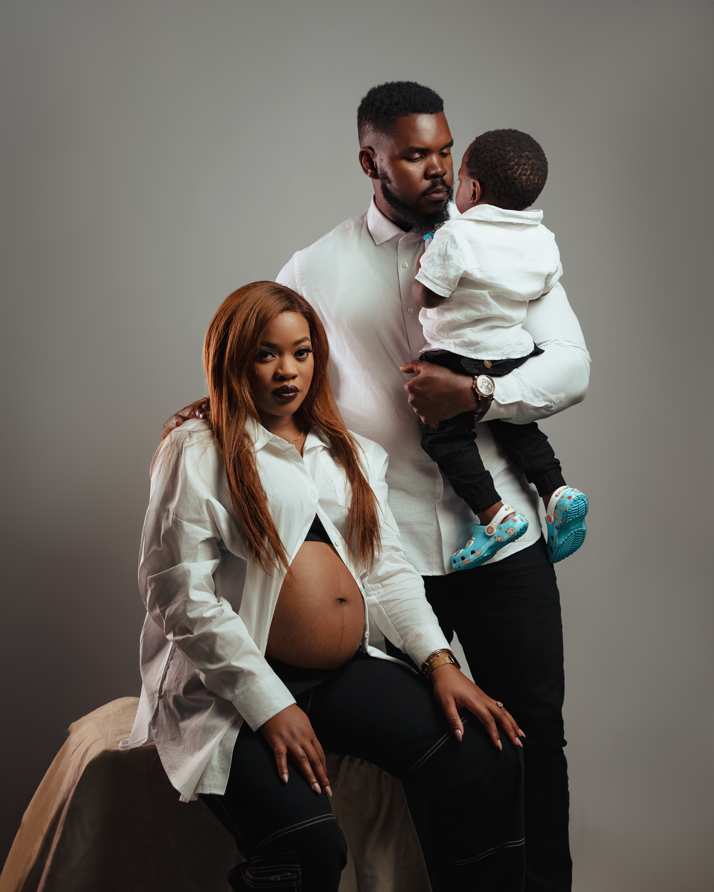 Family maternity photo with white shirts and casual styling against gray background.
