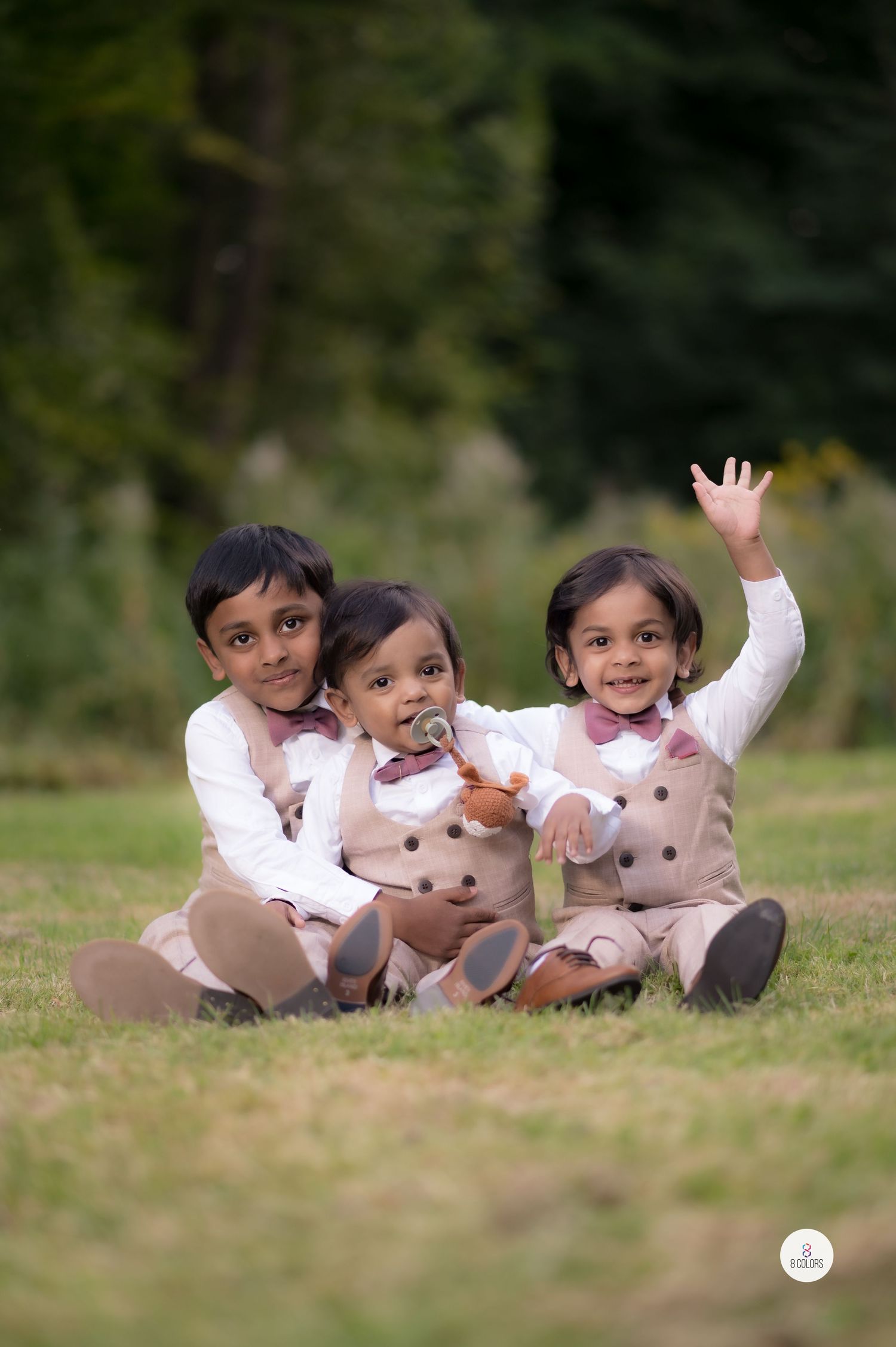 Three children in matching light-colored outfits sit together on grass while posing playfully for a portrait.