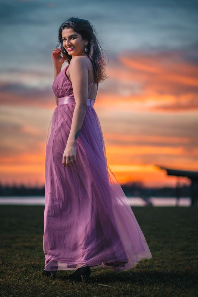 A purple flowing dress catches the evening breeze against a vibrant sunset sky over water.