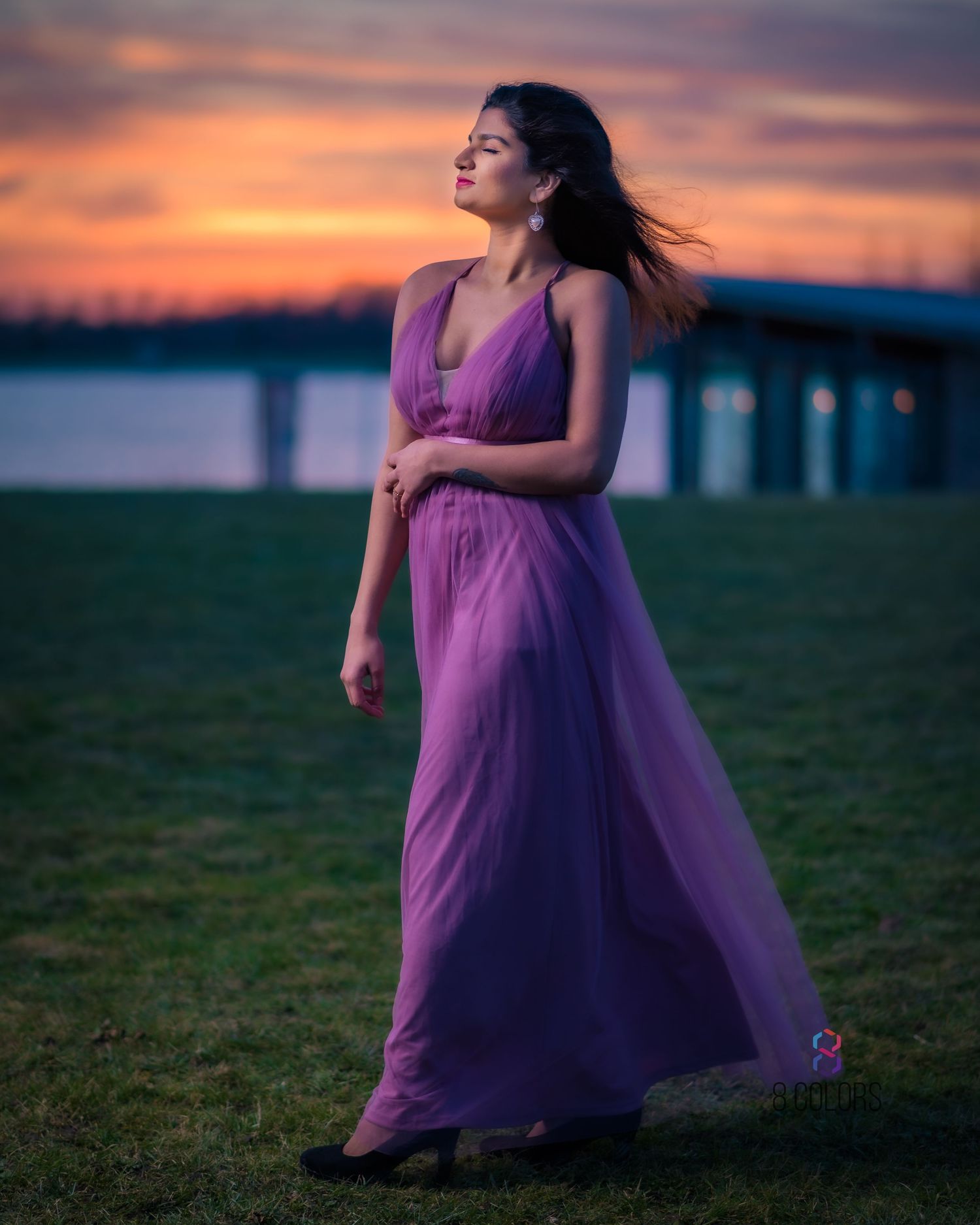 A flowing purple gown catches the breeze against a vibrant sunset sky with a bridge silhouetted in the background.