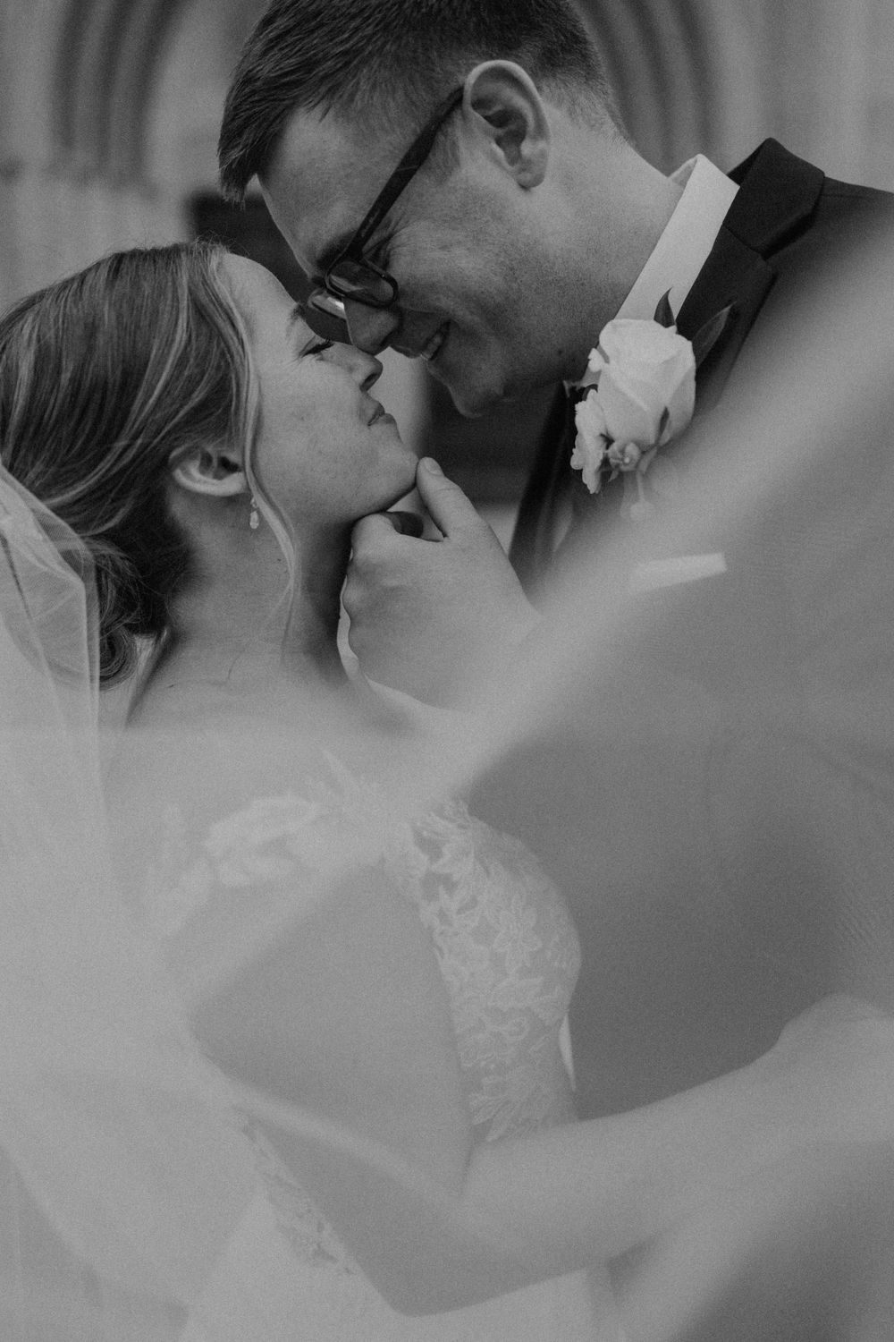 Black and white photo of bride and groom kissing beneath a flowing veil at a church.