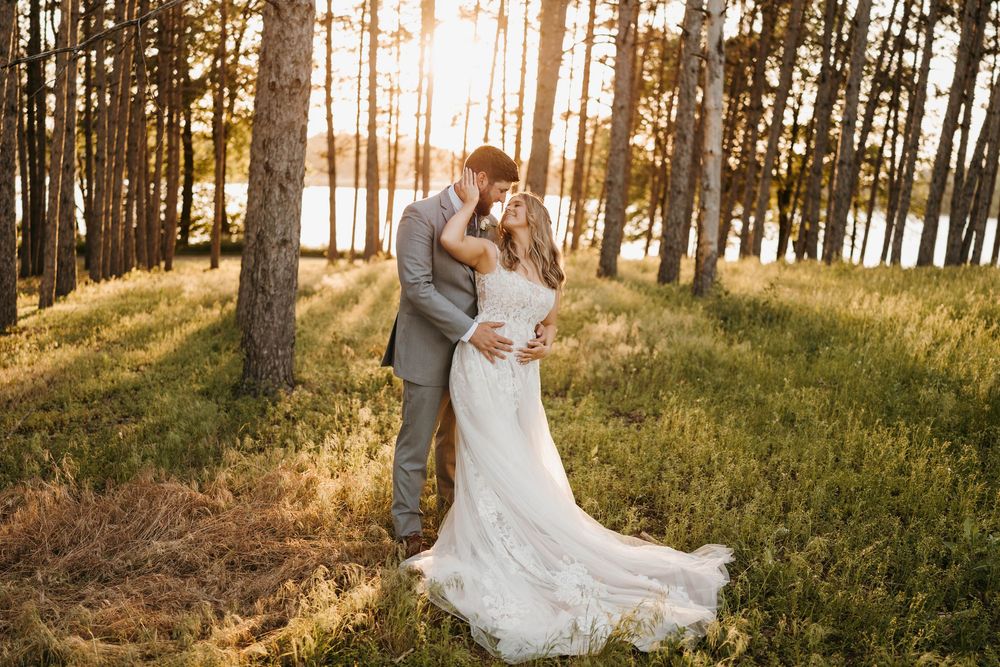 Bride and groom sharing a kiss in sun-drenched pine forest, bride in elegant lace wedding gown.