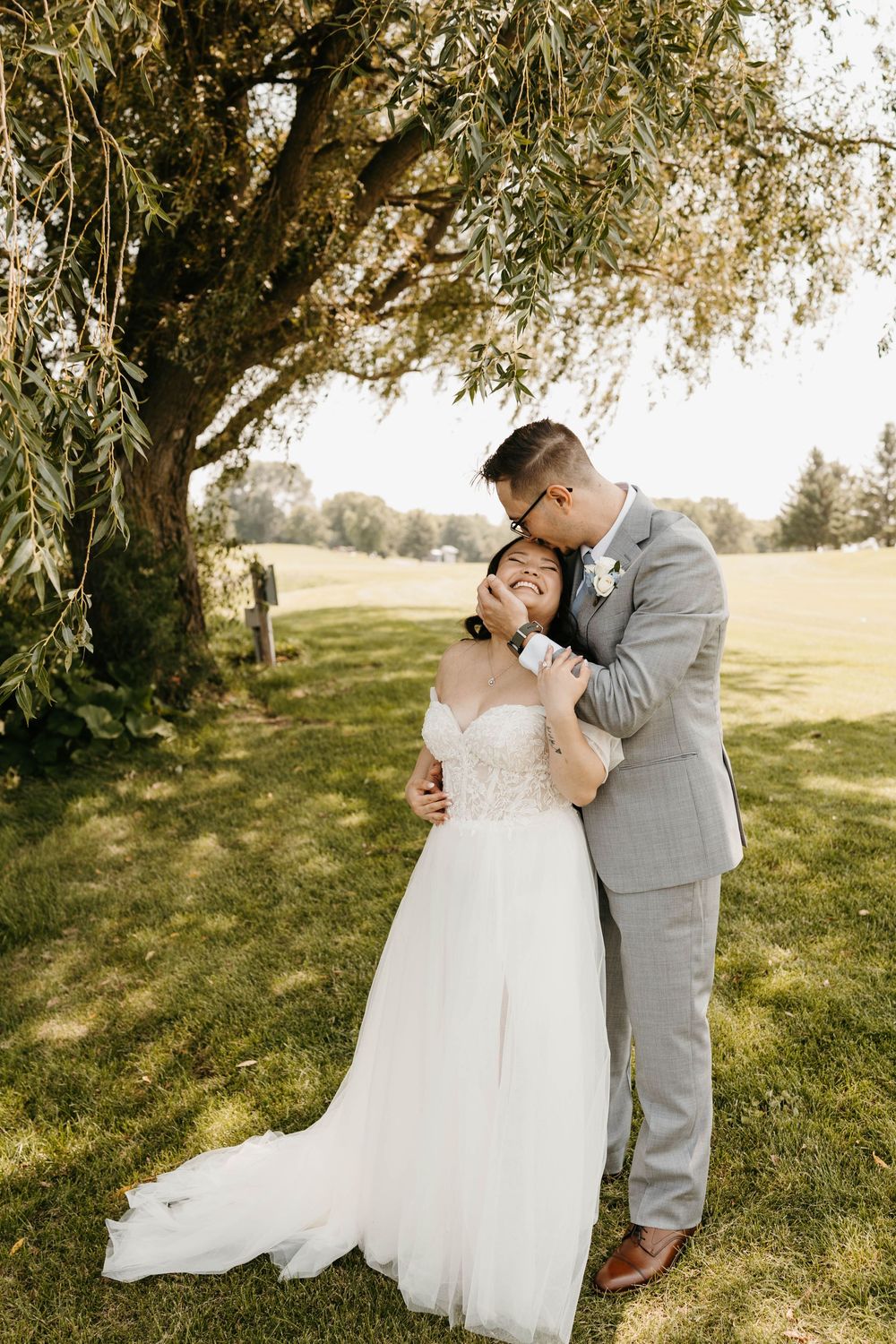 Bride and groom embracing under a willow tree, laughing joyfully, bride in white gown, groom in gray suit, outdoor wedding.