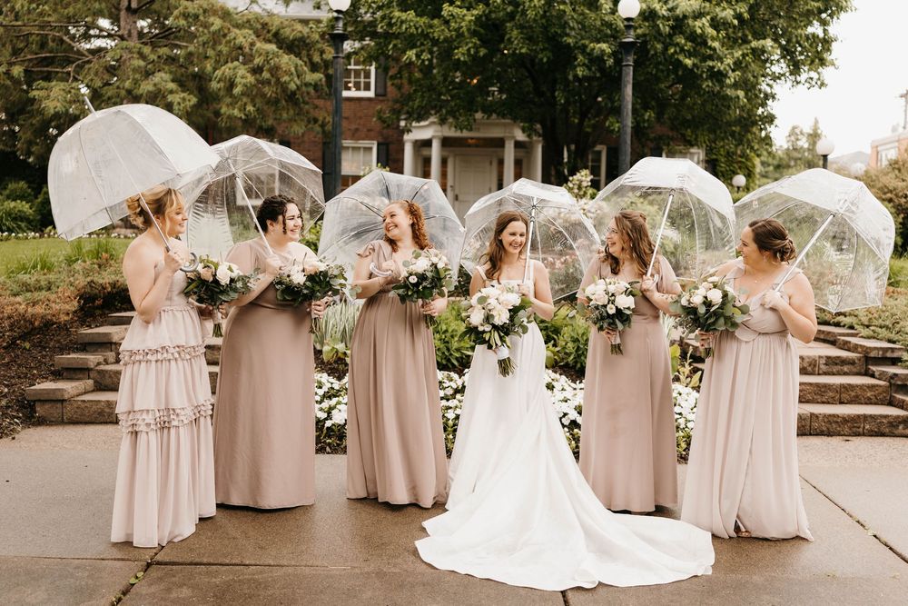 Bride and bridesmaids in blush gowns holding clear umbrellas and white bouquets outdoors.