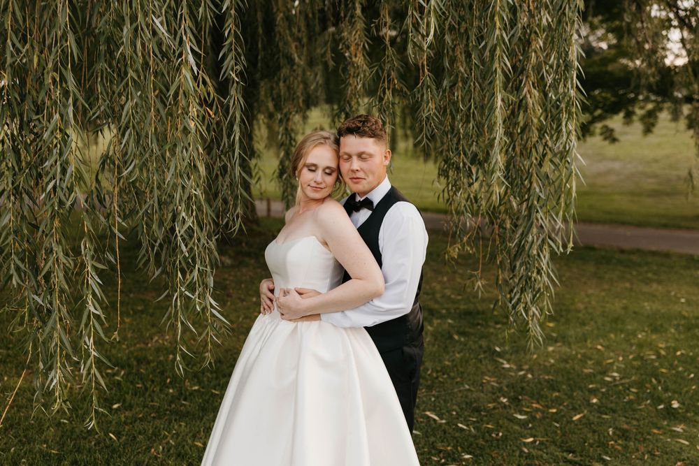 Bride and groom embrace under a weeping willow tree, wearing wedding dress and black bow tie suit.