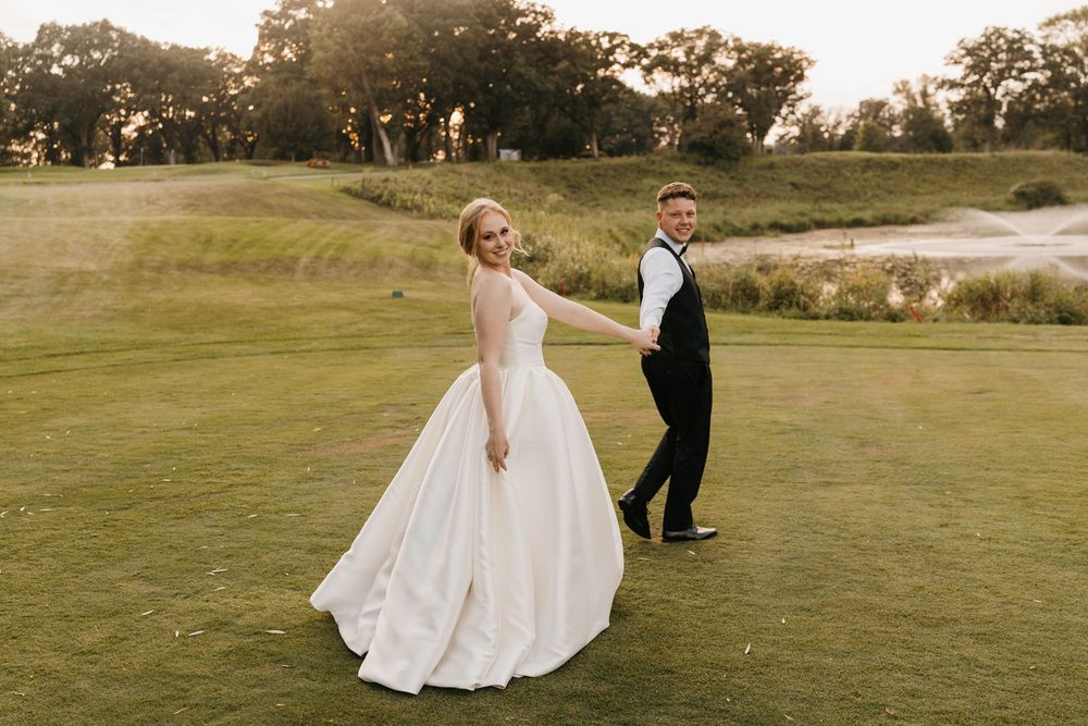 Bride in white ball gown and groom in black suit hold hands on a lush golf course at golden hour.