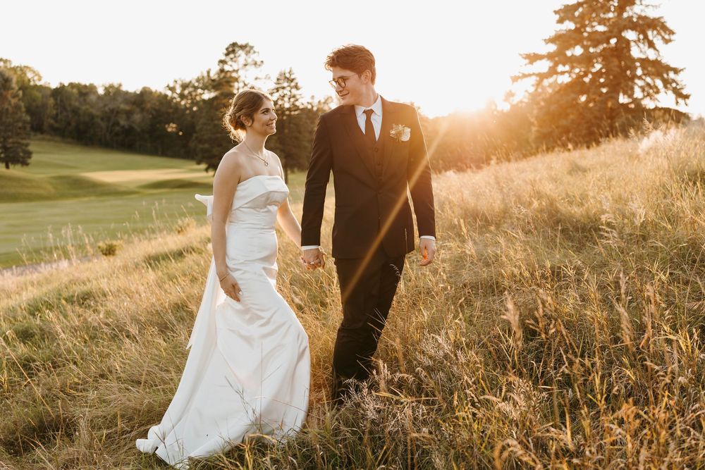 Bride and groom walk hand in hand through golden meadow at sunset in elegant wedding attire.
