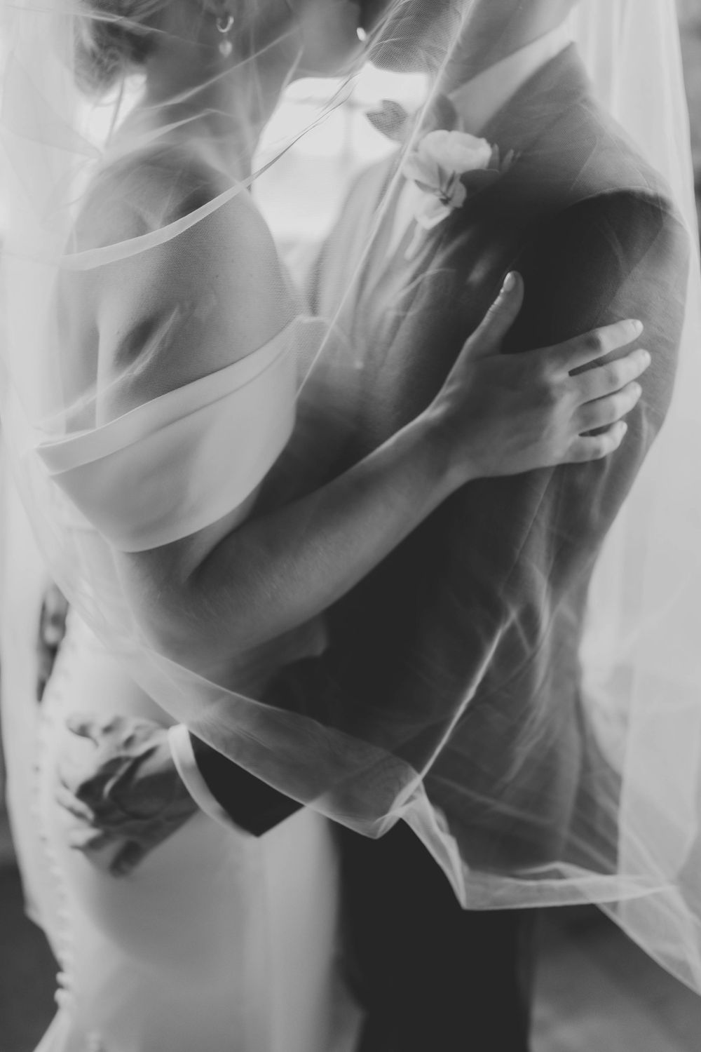 Black and white photo of a bride and groom embracing under a sheer wedding veil.