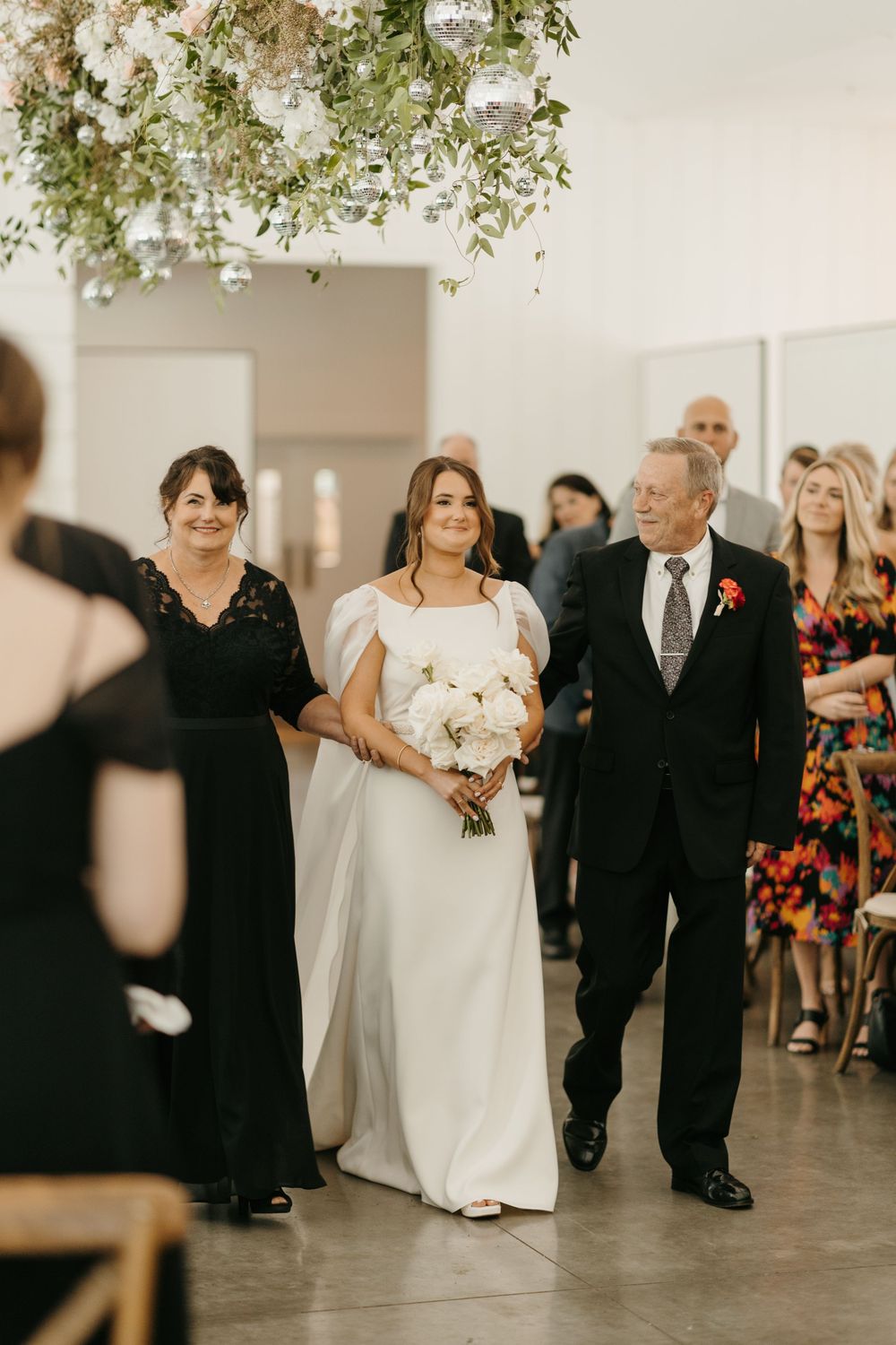 Bride in elegant white gown walks down aisle escorted by parents during wedding ceremony.