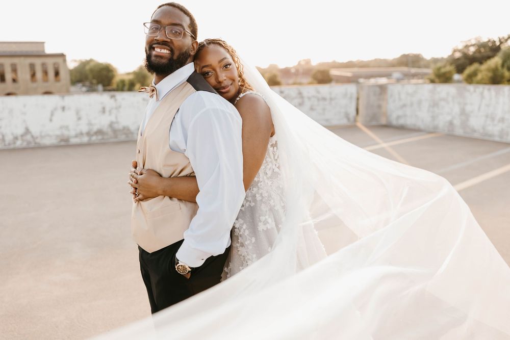 Groom smiles as bride embraces him from behind on rooftop with veil flowing in breeze.