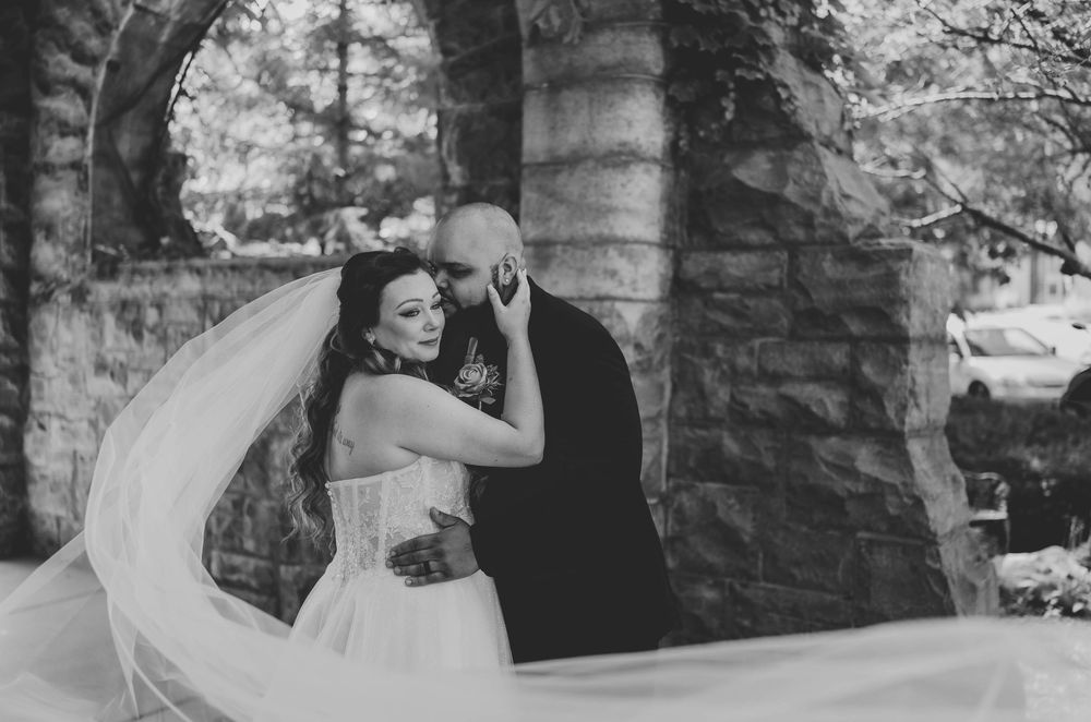 Bride embraces groom near stone arch as her long veil billows dramatically in black and white.