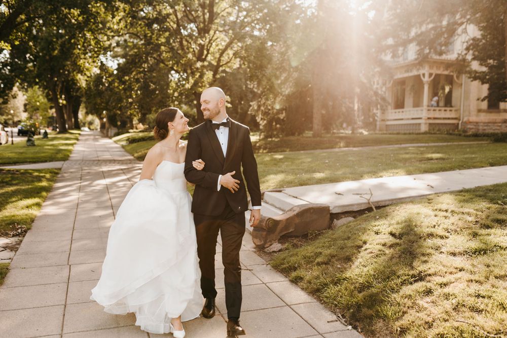 Happy bride and groom walk hand-in-hand down a sunlit sidewalk surrounded by lush green trees.