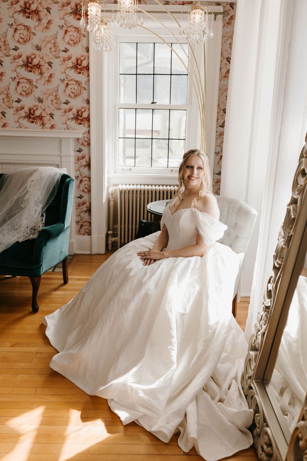 Radiant bride in a voluminous off-shoulder ball gown sits in a sunlit room with floral wallpaper and elegant decor.