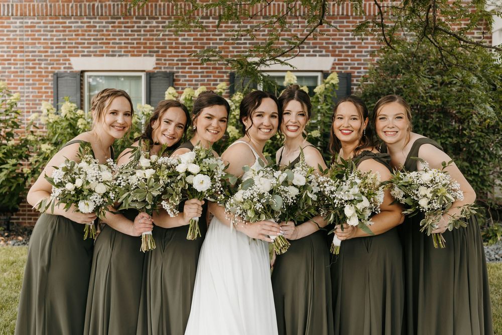 Bride in white gown with five bridesmaids in olive green dresses holding white floral bouquets outdoors.