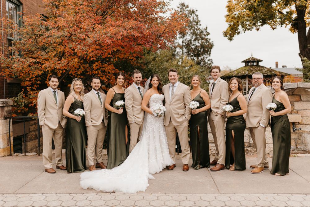 Wedding party posing outdoors in tan suits and olive green dresses with vibrant fall foliage backdrop.