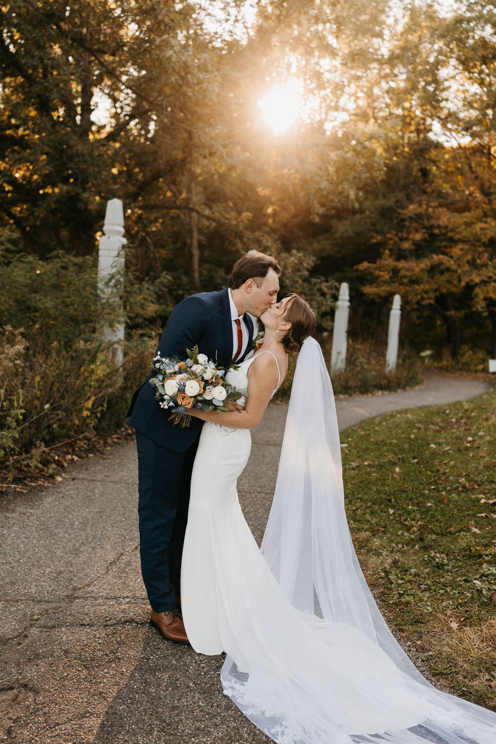 Bride and groom share a romantic kiss at sunset in a garden, her long veil trailing behind as she holds a floral bouquet.