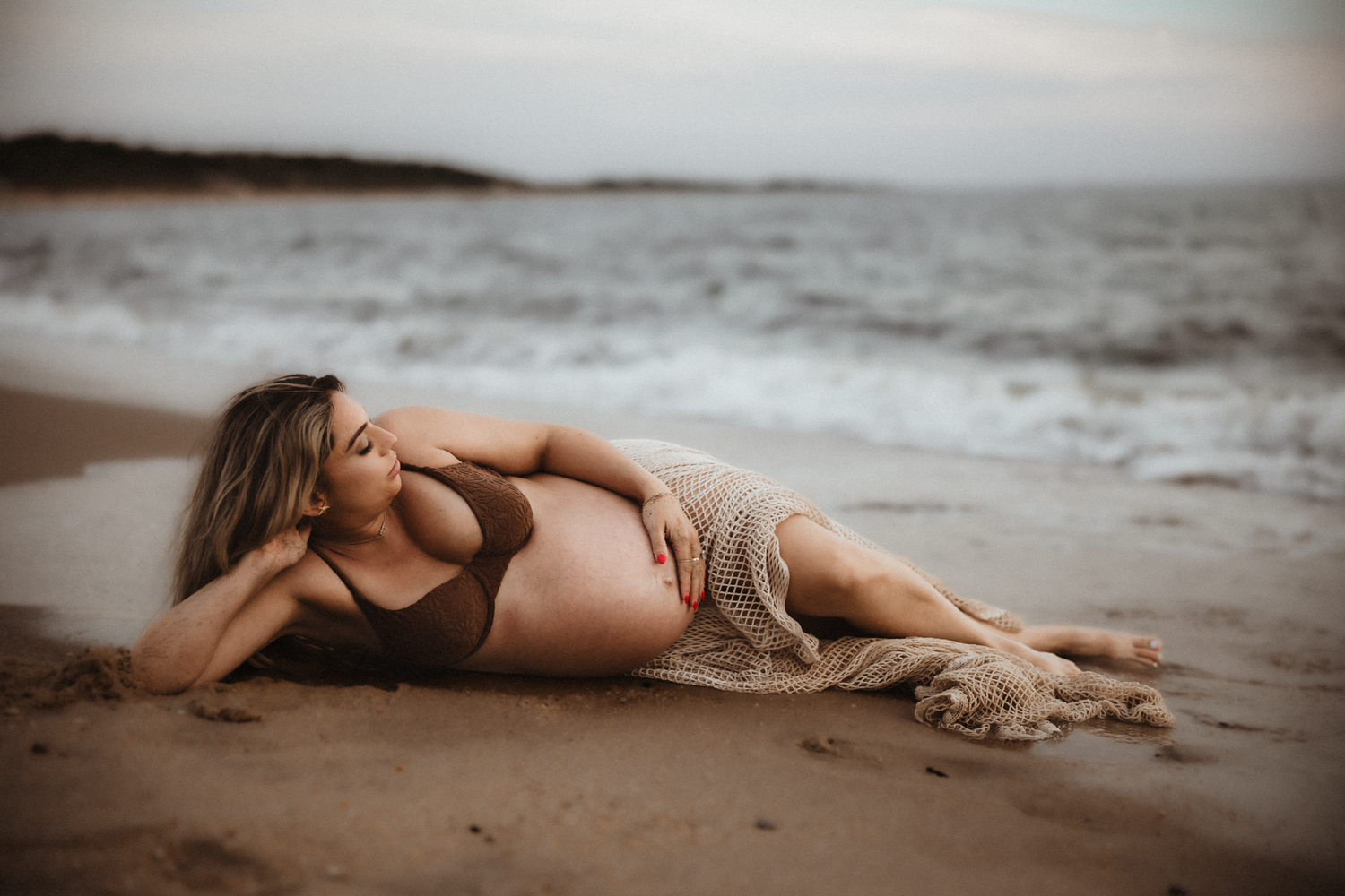 Artistic maternity photo of woman in brown dress lying in shallow water at sunset.