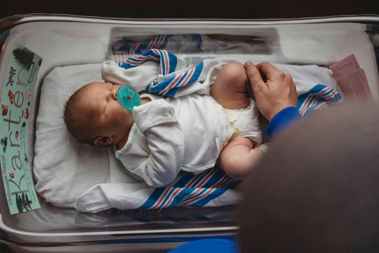 A newborn baby rests in a hospital bassinet wrapped in blue and white striped blanket.