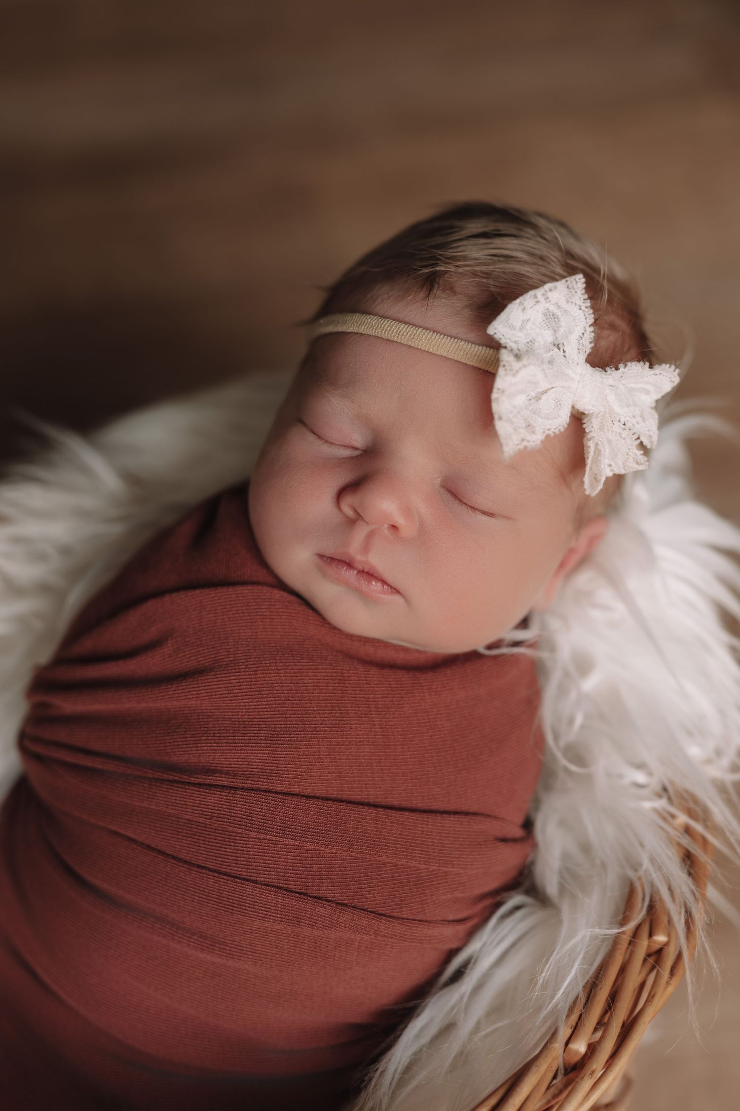 Sleeping newborn wrapped in beige swaddle with floral bow headband on white fur.