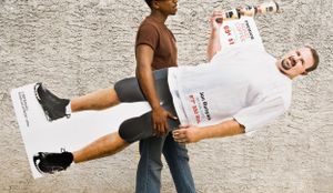 A Black man carries a White man holding a fitness advertisement against a concrete wall.