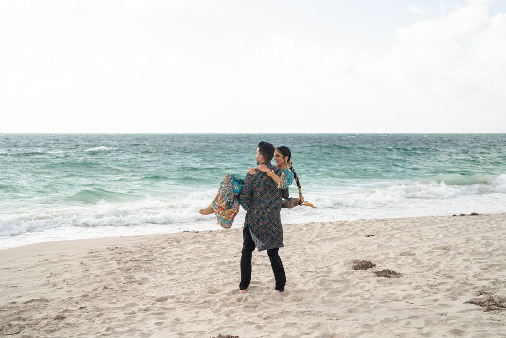 Romantic beach moment with groom carrying bride along shoreline at sunset.
