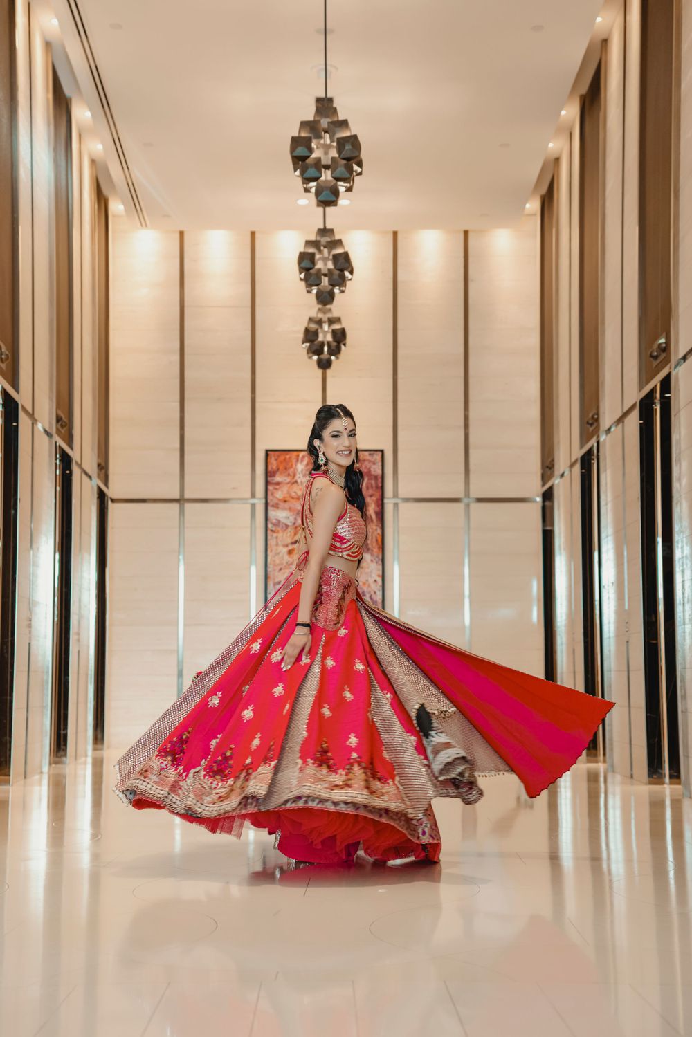 Bride in stunning red lehenga twirling in elegant hotel hallway with modern chandeliers.