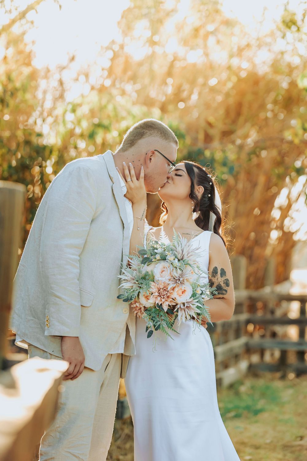 Newlywed couple shares romantic kiss in sunlit outdoor setting with rustic fence and autumn foliage backdrop.