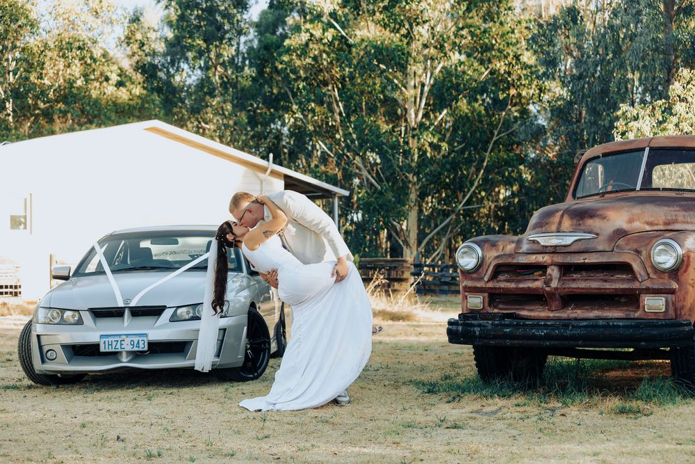 A couple shares a romantic moment in a rustic setting between a modern white car and vintage brown pickup truck.