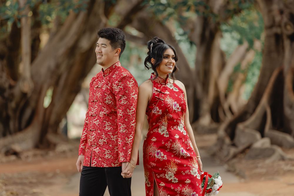 A couple wearing matching red Chinese traditional outfits pose in a wooded outdoor setting.