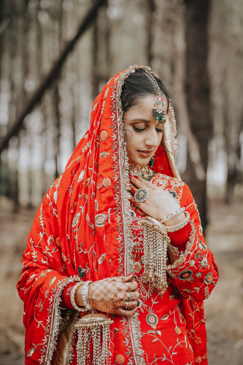 Bride in ornate red Indian wedding attire with detailed embroidery and jewelry poses in forest setting.