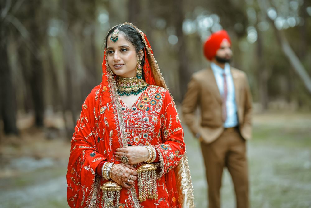 Traditional Indian bride in red lehenga poses in forest while groom in brown suit stands in background.