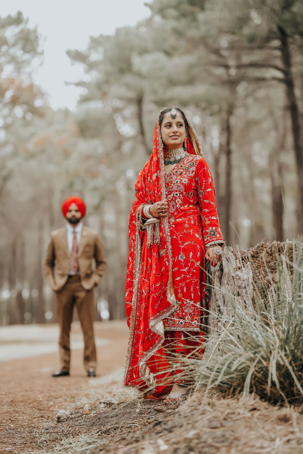 Bride in red traditional Indian wedding dress poses on forest path with groom in distance.
