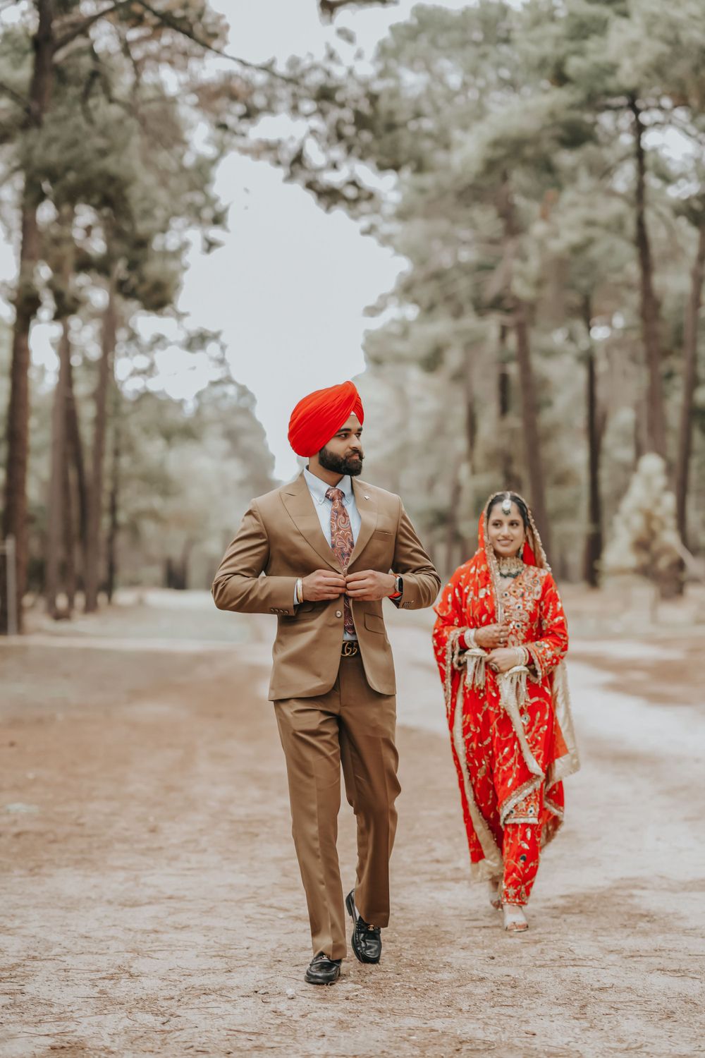 Sikh couple in traditional wedding attire walk along forest path during wedding photoshoot.