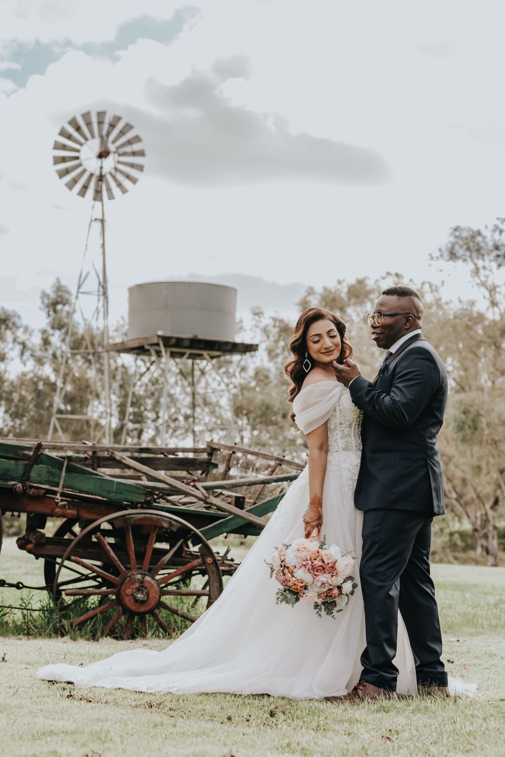 Romantic wedding portrait with rustic windmill and vintage wagon in countryside setting.