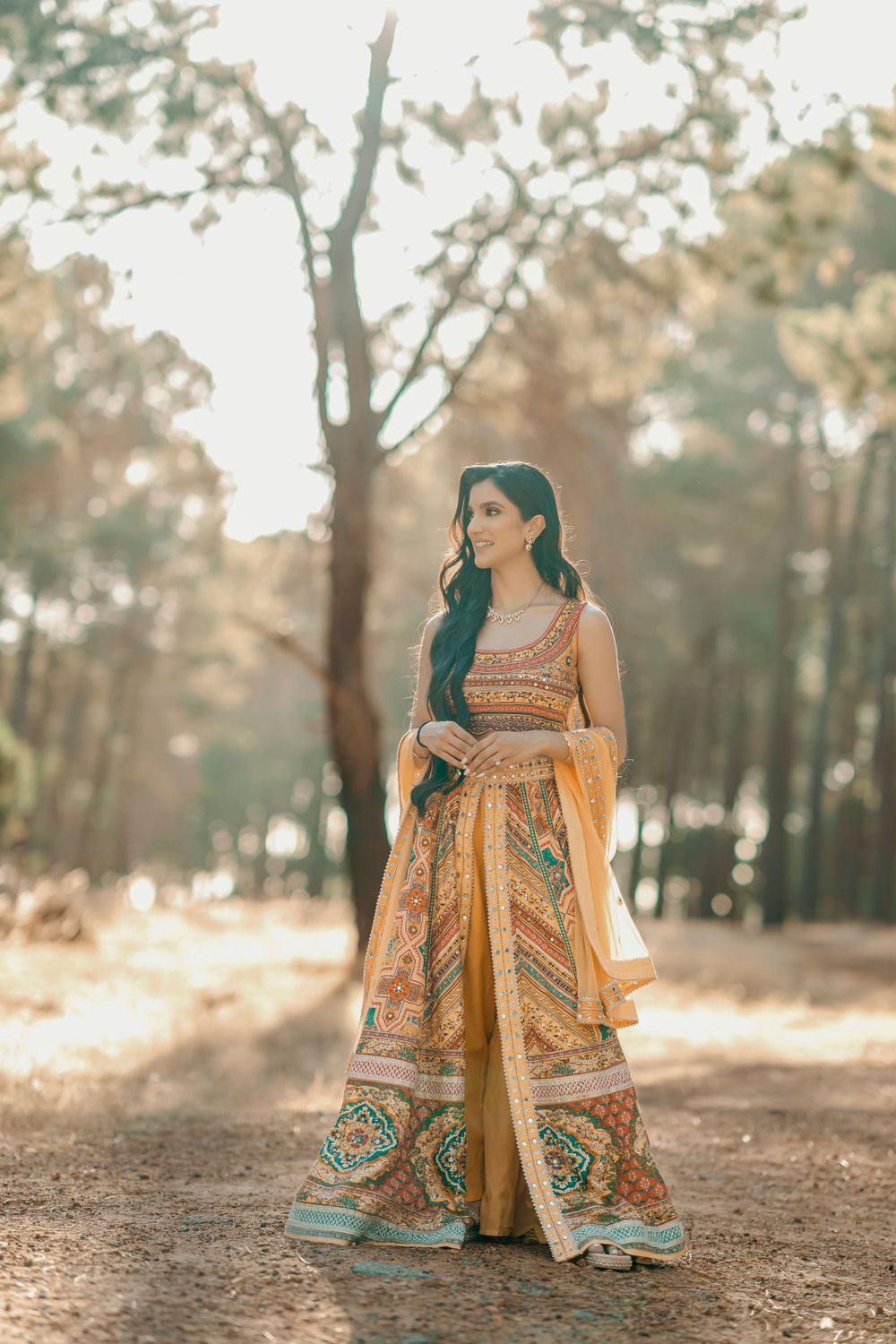 Woman in ornate traditional Indian wedding attire posing in sunlit forest setting.