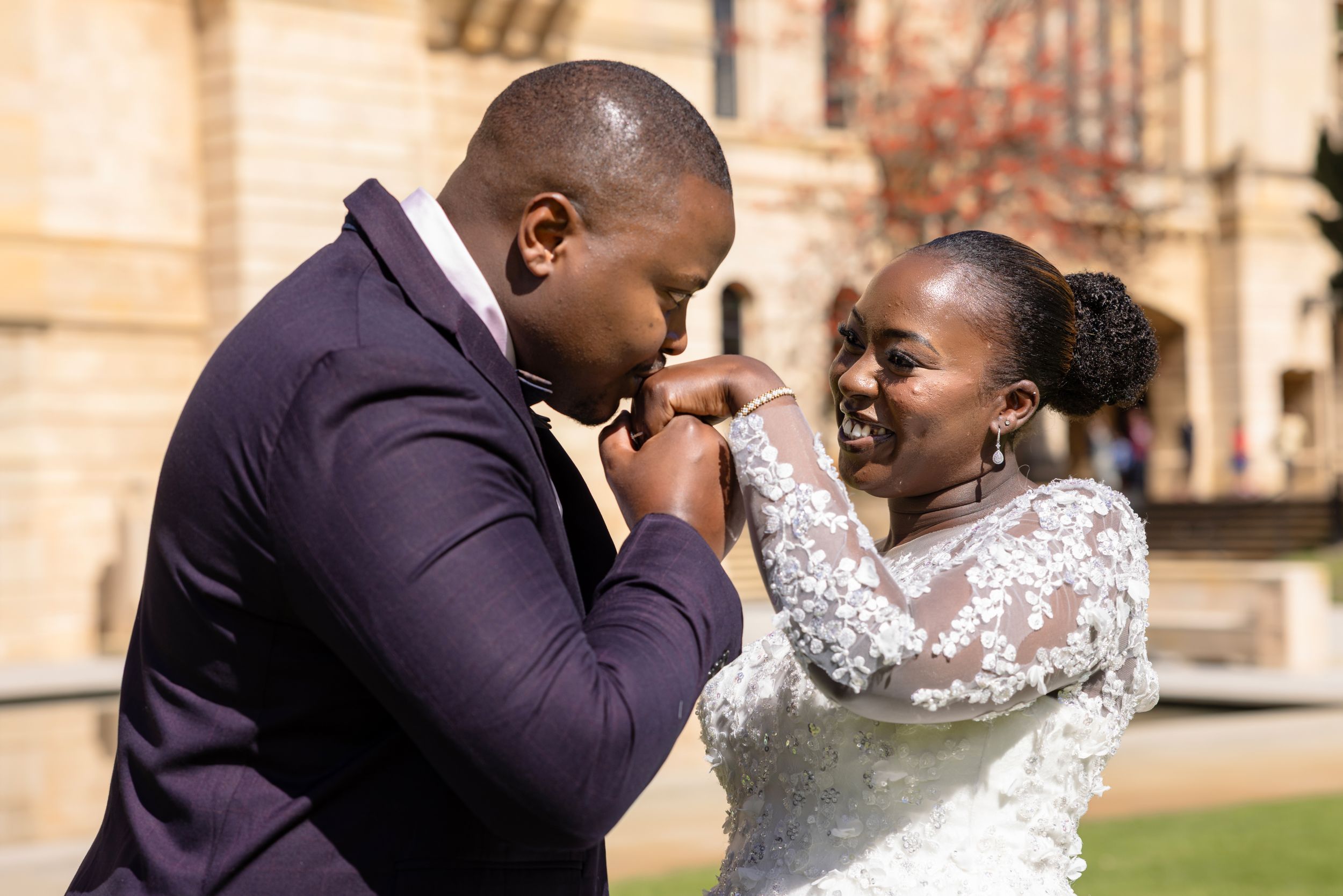 A happy couple shares multiple romantic kisses outside a rustic brick building on a sunny day.
