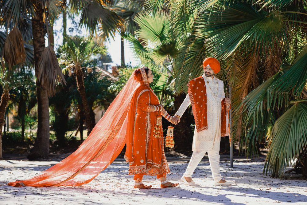 Couple in orange and white Indian wedding outfits walk together in tropical garden setting.