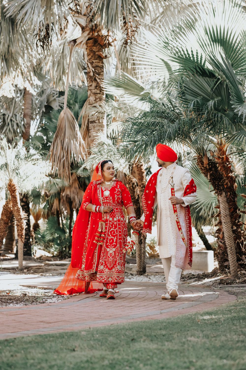 Wedding couple in traditional Indian attire pose among palm trees in garden setting.