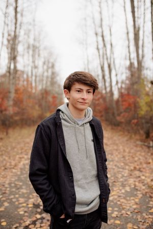 A person wearing a black jacket and grey hoodie stands on a path covered in autumn leaves.