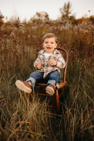 A toddler sits in wooden chair among tall grass wearing plaid and jeans.