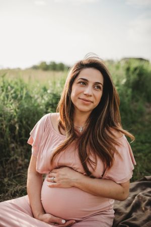 Expecting mother in pink dress sitting outdoors during golden hour maternity shoot.