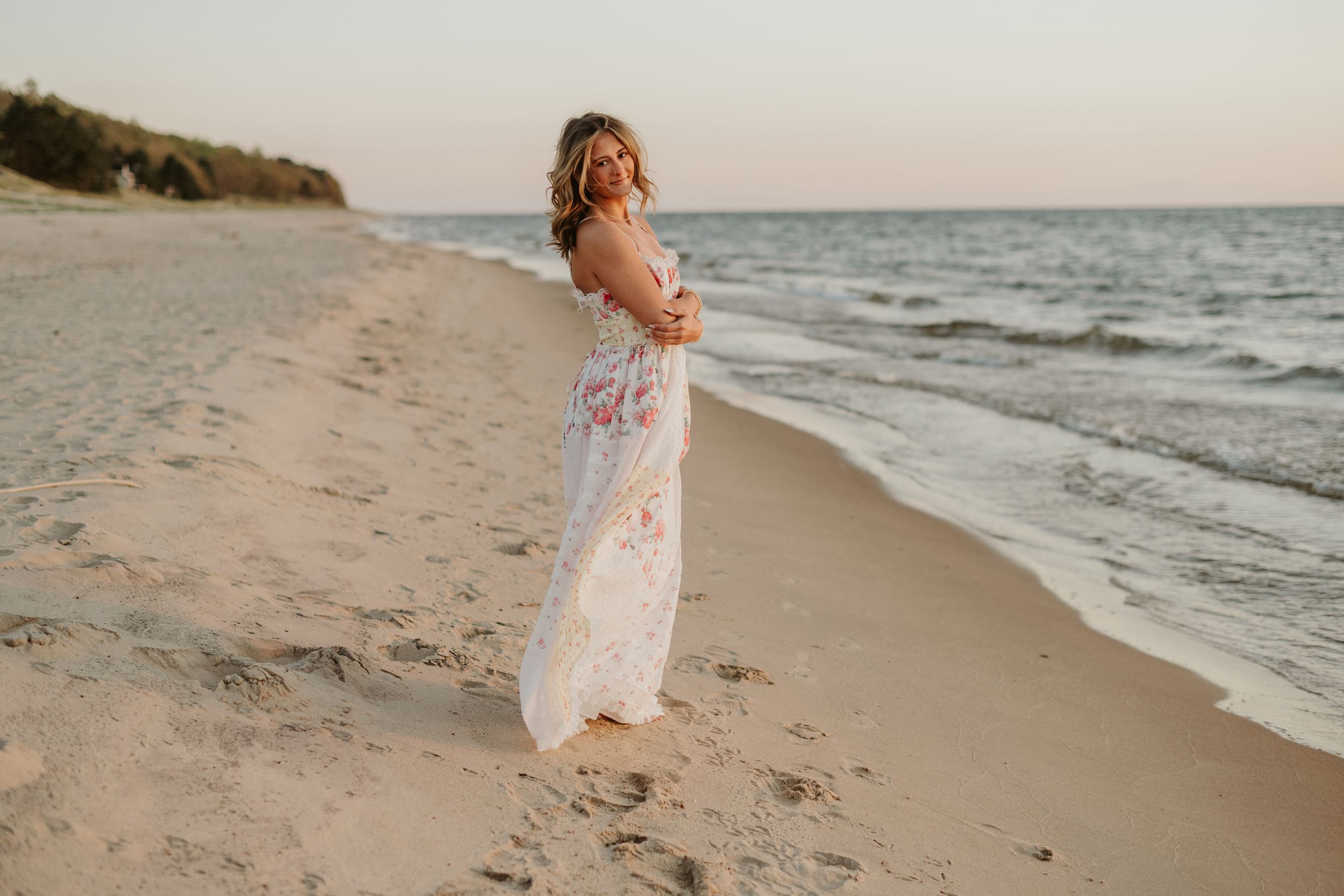 Pregnant woman in floral maxi dress standing on sandy beach at sunset, smiling over shoulder near ocean waves.