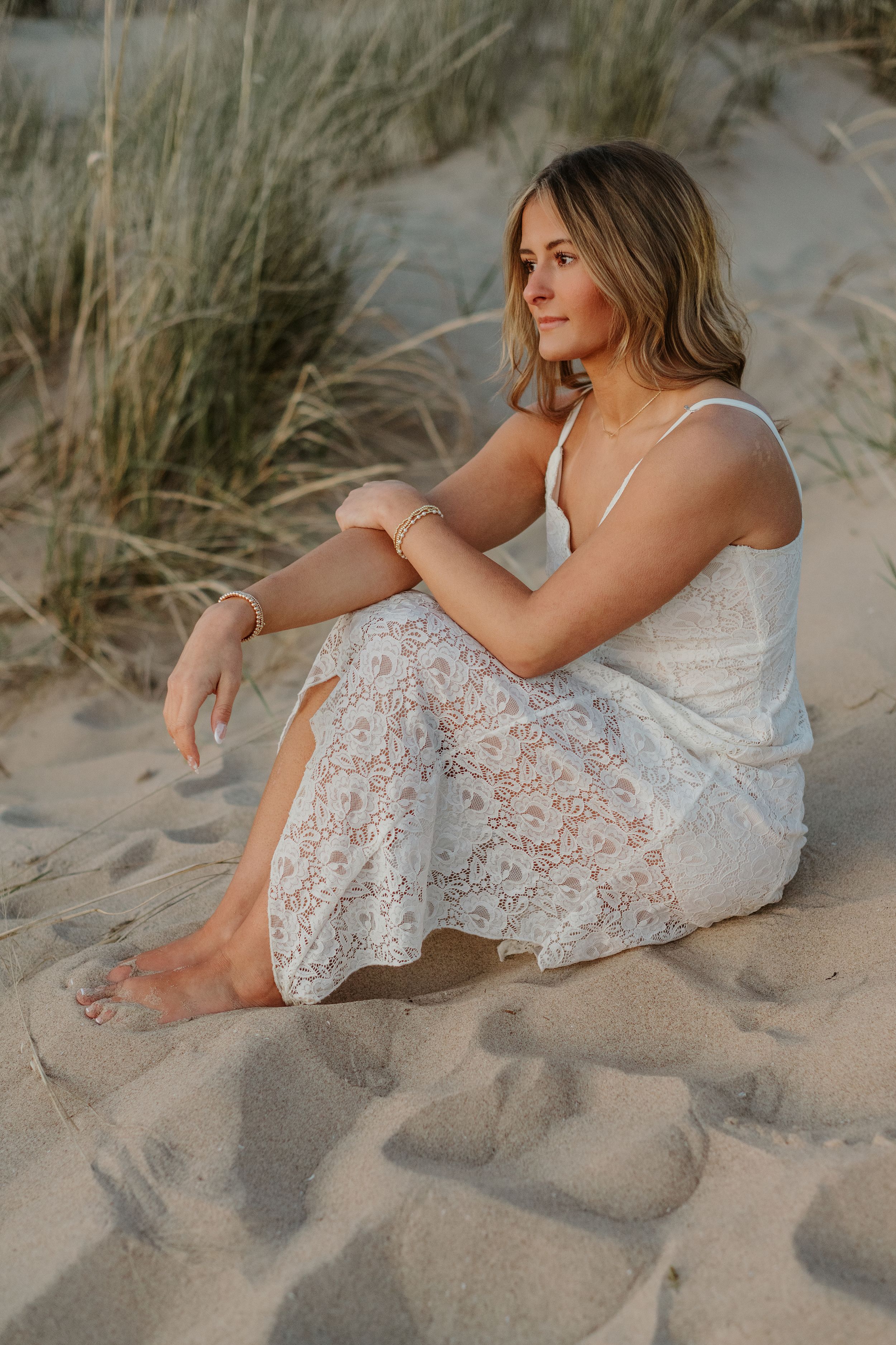 Young woman in white lace dress sitting barefoot on sandy beach dunes at golden hour.