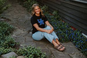 Smiling young woman in University of Kentucky shirt sitting on stone path surrounded by blue flowers.