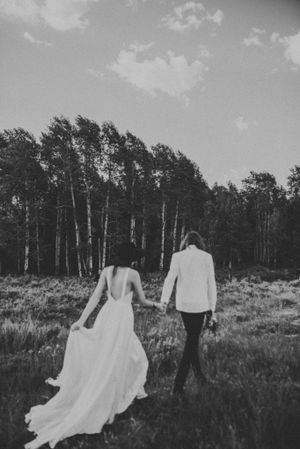 Black and white photo of couple walking hand in hand through forest field wearing wedding attire.