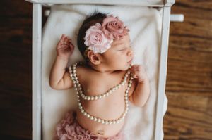 Sleeping newborn wearing pearl necklaces and pink floral headband in white wooden box.