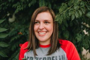 A person in a red baseball shirt smiles against a leafy green background.